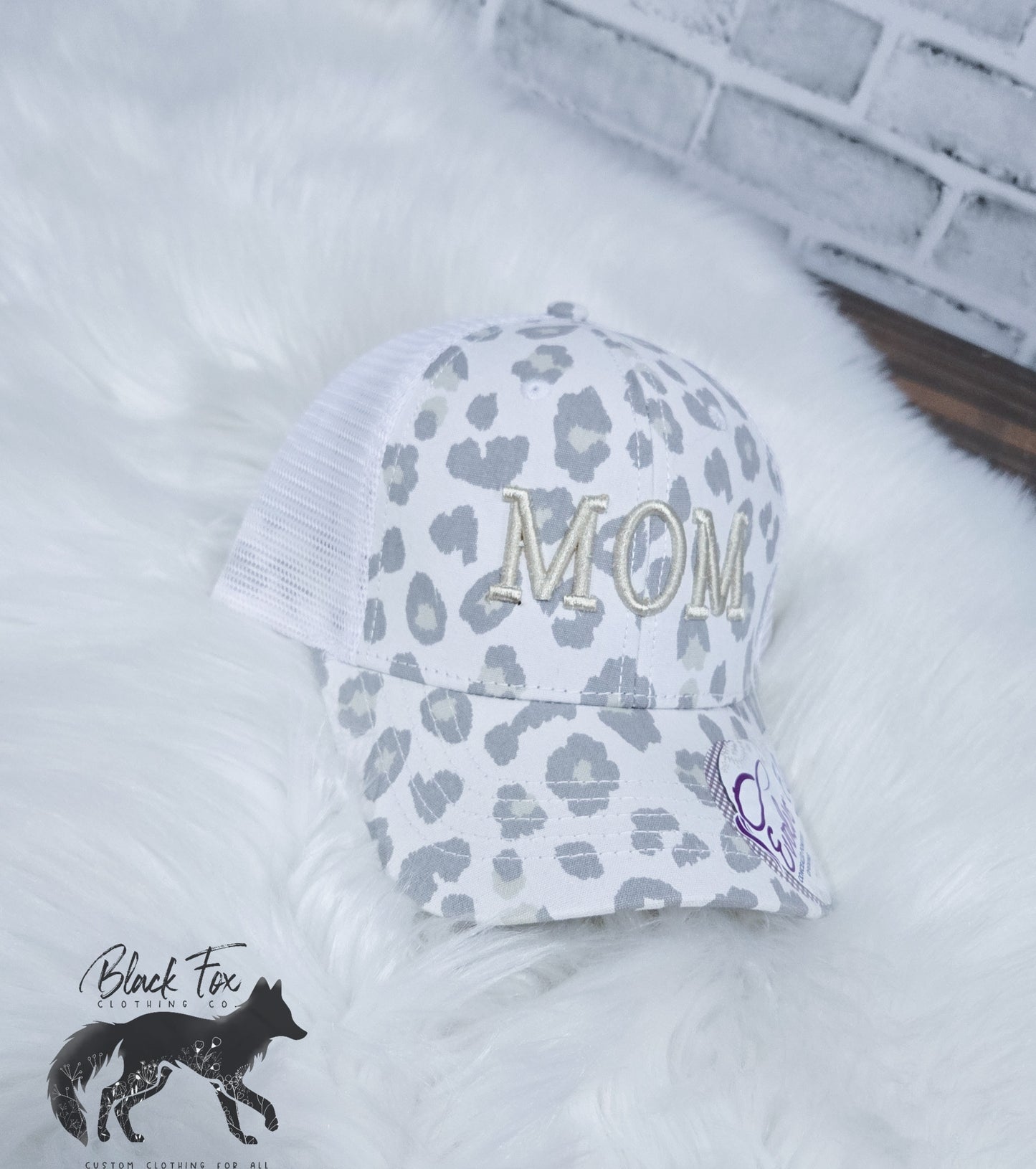 A trucker hat with a cream and brown leopard print and the letters 'MOM' embroidered in gold on the front. The hat is displayed on a white furry surface.