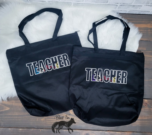 Two black tote bags with 'TEACHER' printed on them, placed on a textured surface.