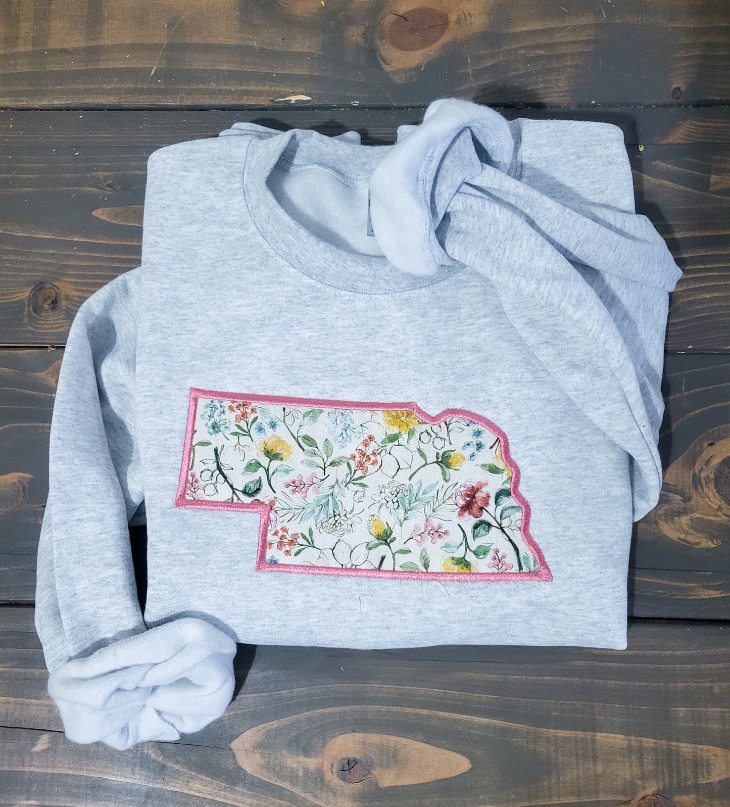 A gray crewneck sweatshirt with a floral state applique design laid out on a wooden surface.