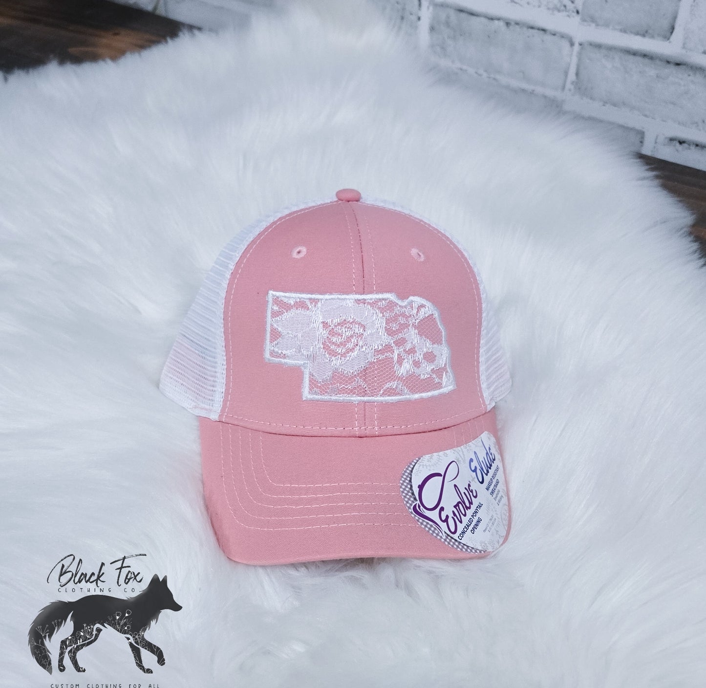 A pink Nebraska Lace applique Trucker Hat displayed on a white surface, featuring a white mesh back and a state outline design on the front.