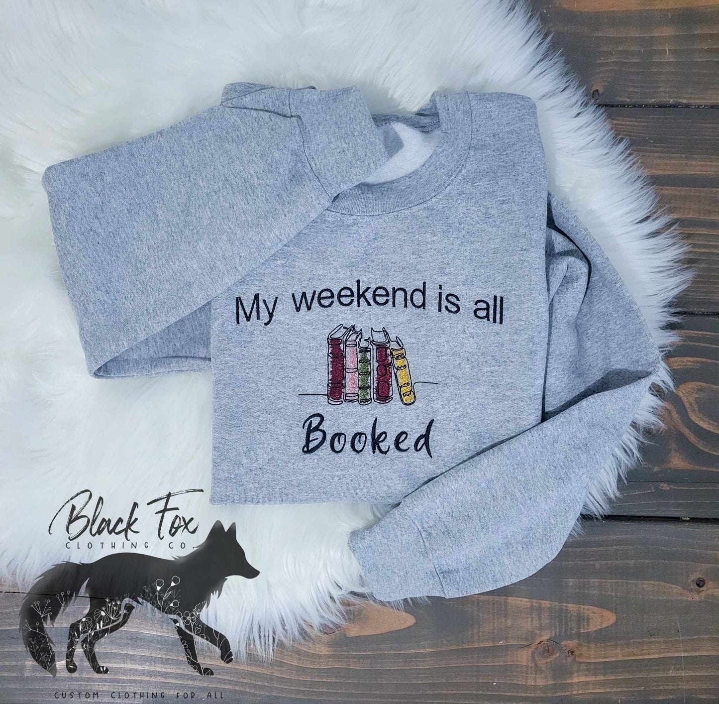 A gray crewneck sweatshirt with the phrase 'My weekend is all booked' embroidered on the front, displayed on a white fluffy surface with a wooden background. A black fox silhouette is also visible.