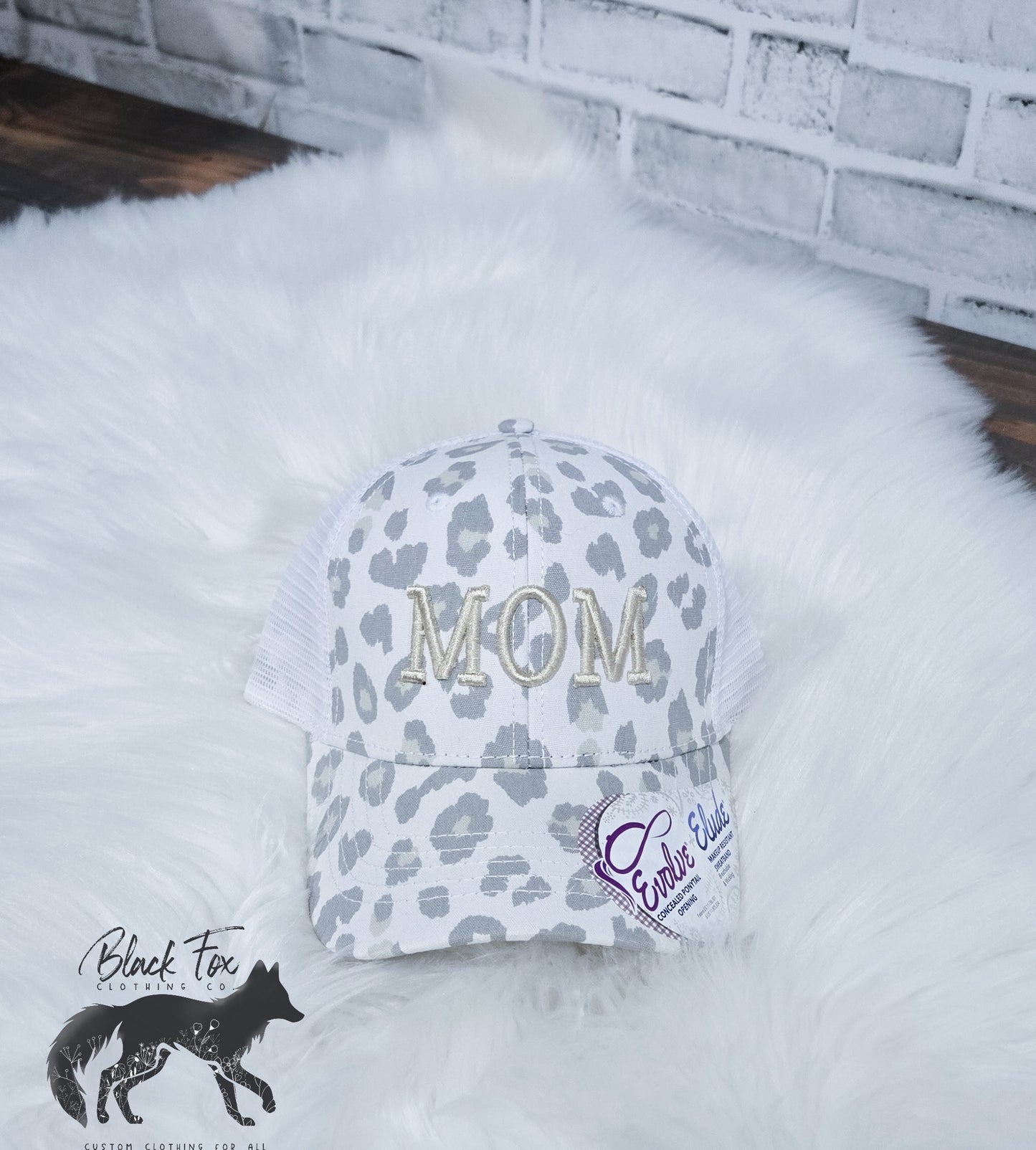 A white leopard print trucker hat with 'MOM' embroidered in capital letters on the front.