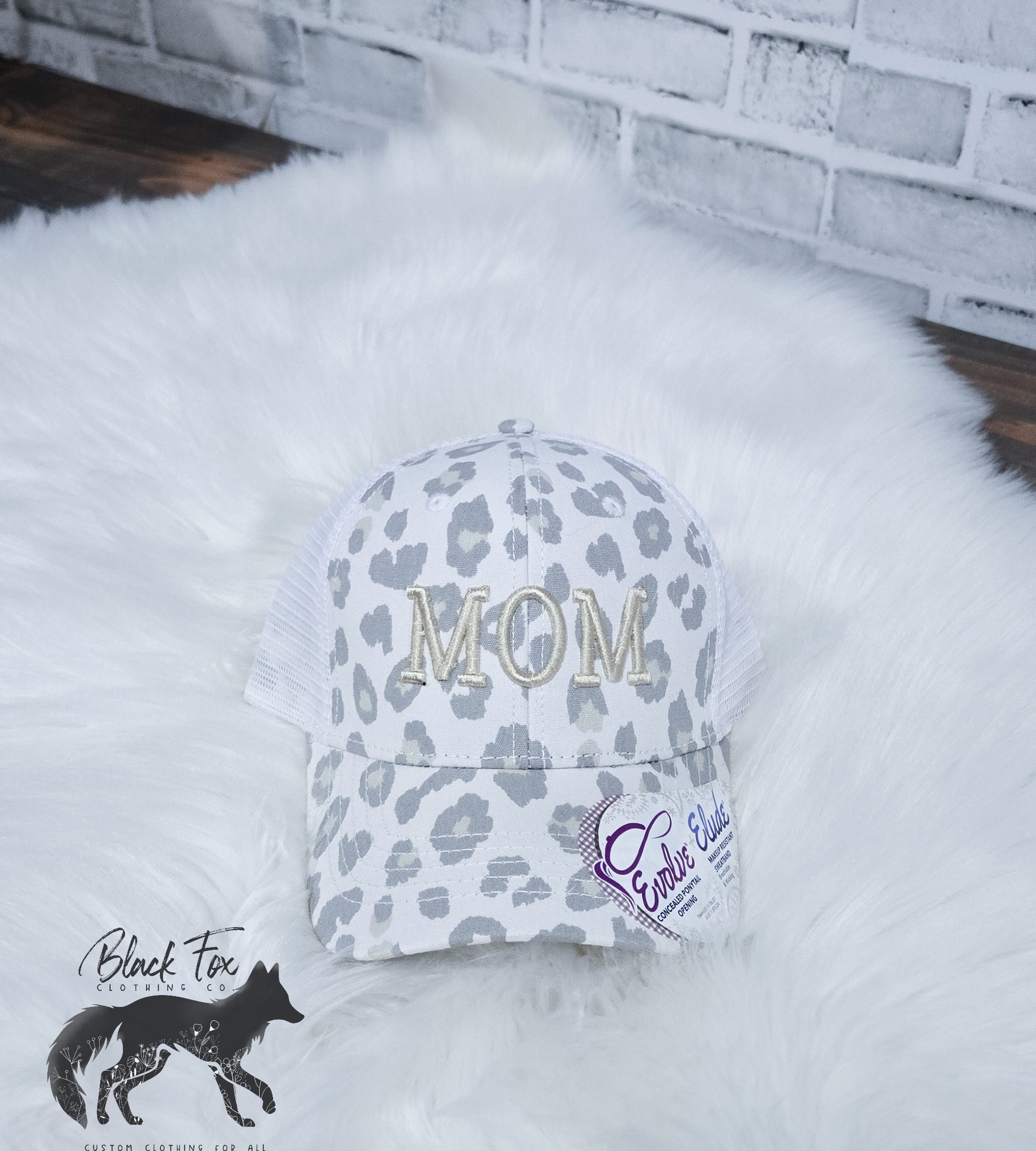 A white leopard print trucker hat with 'MOM' embroidered in capital letters on the front.