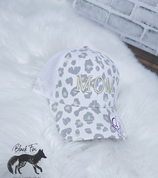 A trucker hat with a cream and brown leopard print and the letters 'MOM' embroidered in gold on the front. The hat is displayed on a white furry surface.