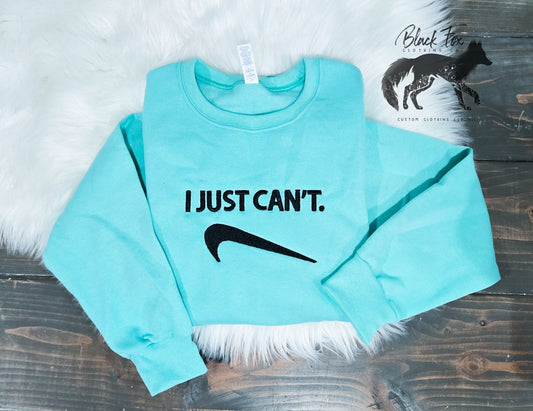 A blue gildan crewneck sweatshirt with the text 'I JUST CAN'T.' followed by a graphic of a nail, laid out flat on a wooden surface with a white fluffy object to the left.