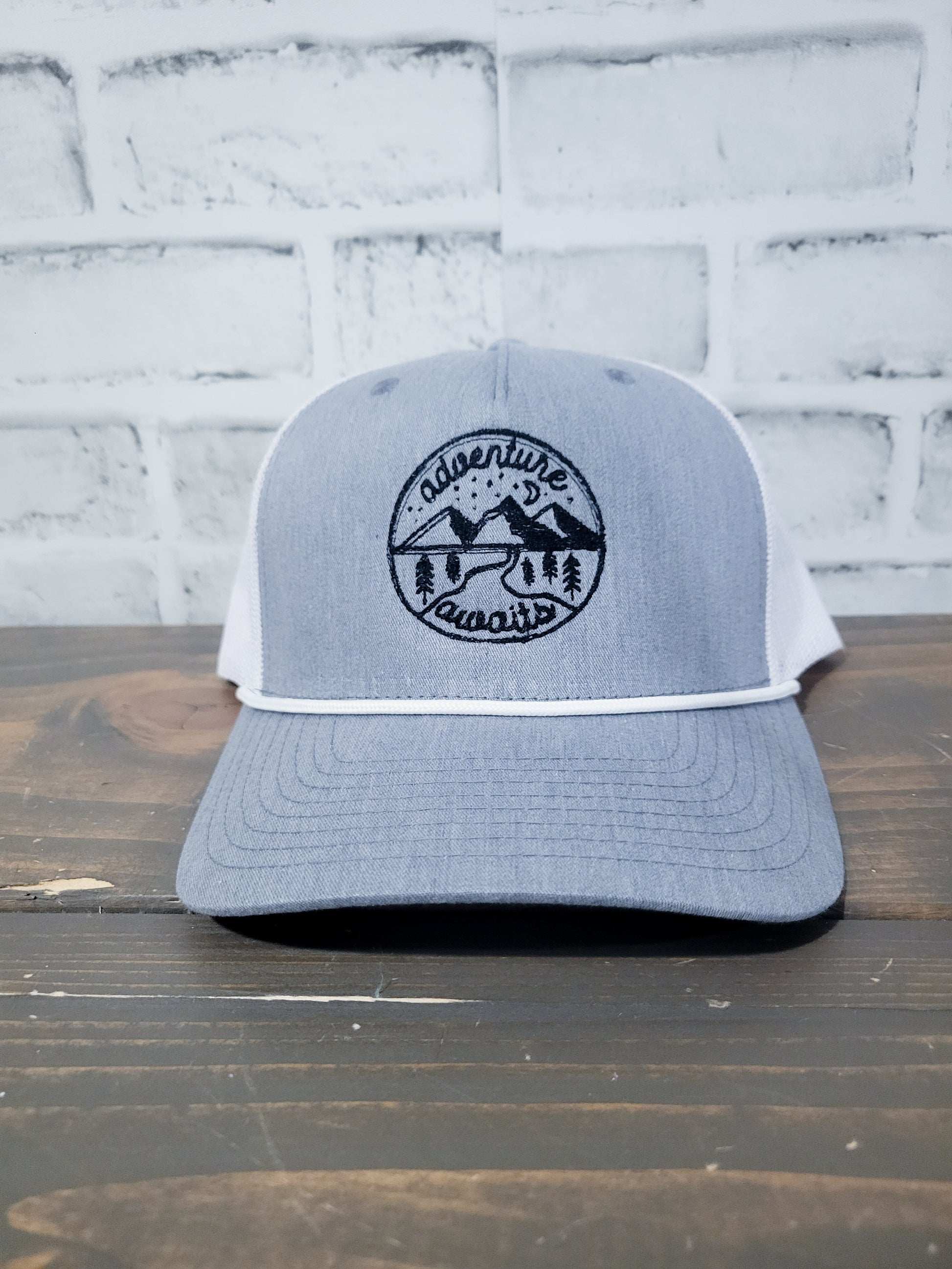 a black circle with mountains and a trees with adventure arched across the top and awaits arched around the bottom of a gray trucker hat.