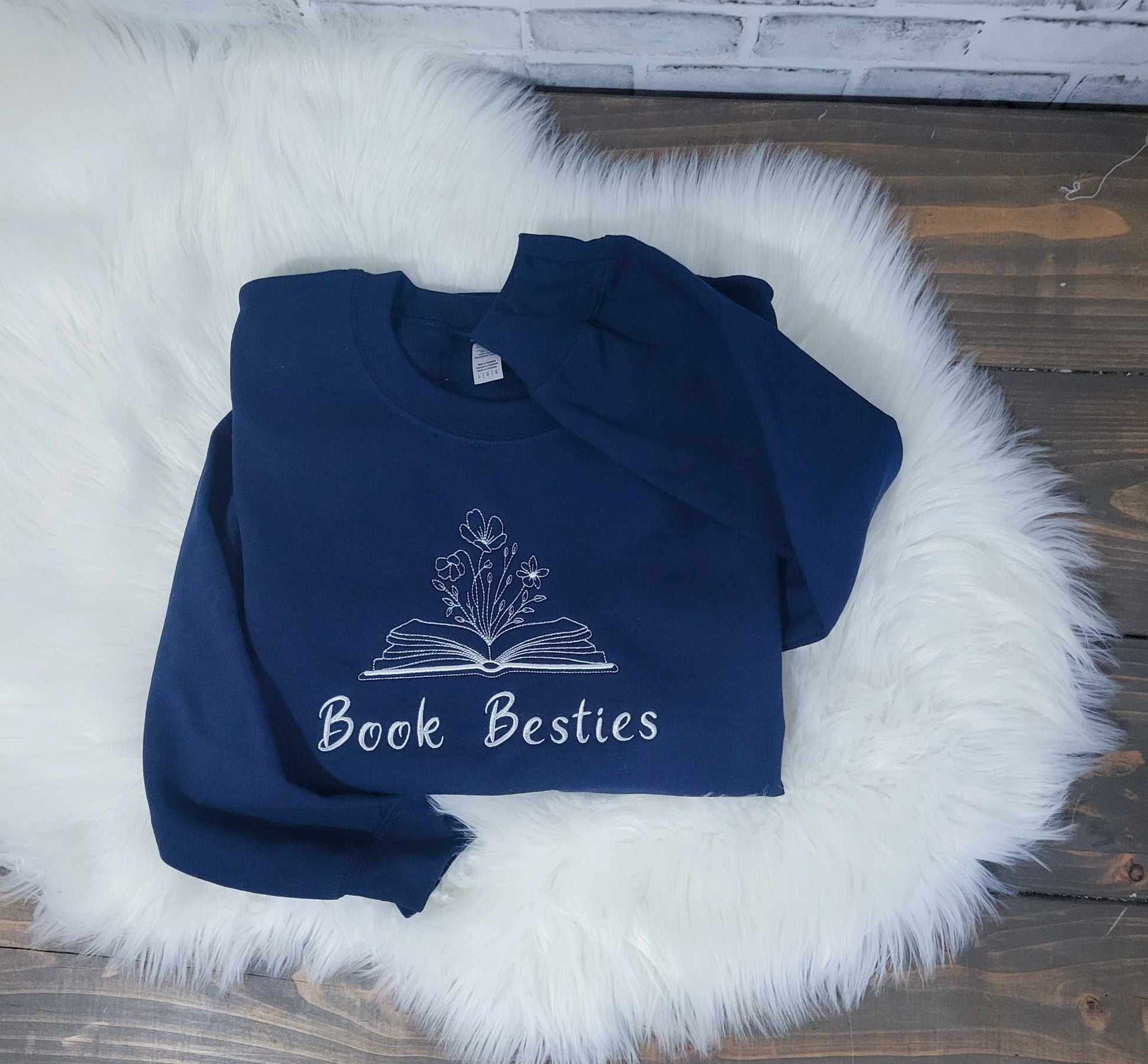 A navy blue crewneck sweatshirt with embroidered text 'Book Besties' laid flat on a white furry surface.