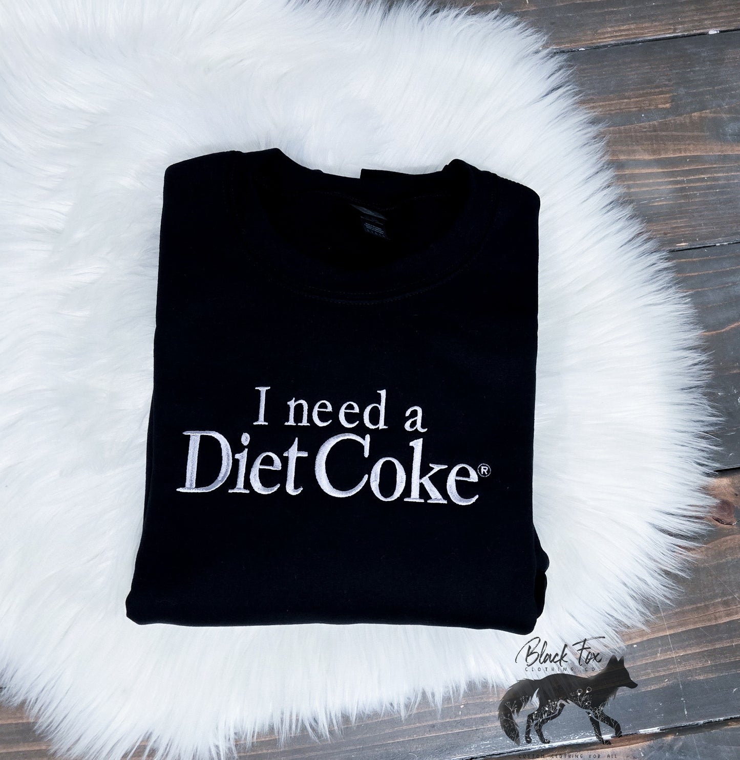 Black fleece crewneck sweatshirt with the text 'I need a Diet Coke' printed on the front, displayed on a white fluffy surface.