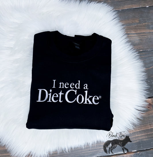 Black fleece crewneck sweatshirt with the text 'I need a Diet Coke' printed on the front, displayed on a white fluffy surface.