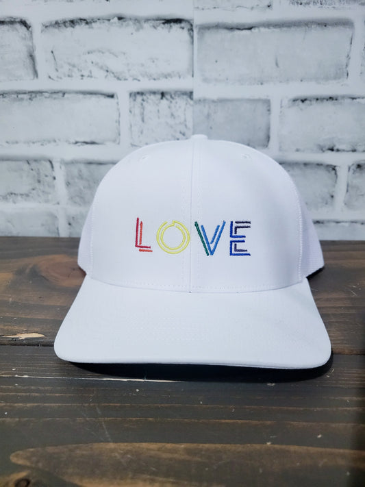 2d word LOVE in red, orange, yellow, green, blue, indigo and violet letters on a white trucker hat