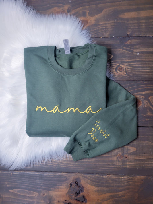A green crewneck sweatshirt laid flat on a wooden surface with 'mama' written in gold script on the front and a name embroidered on the sleeve.