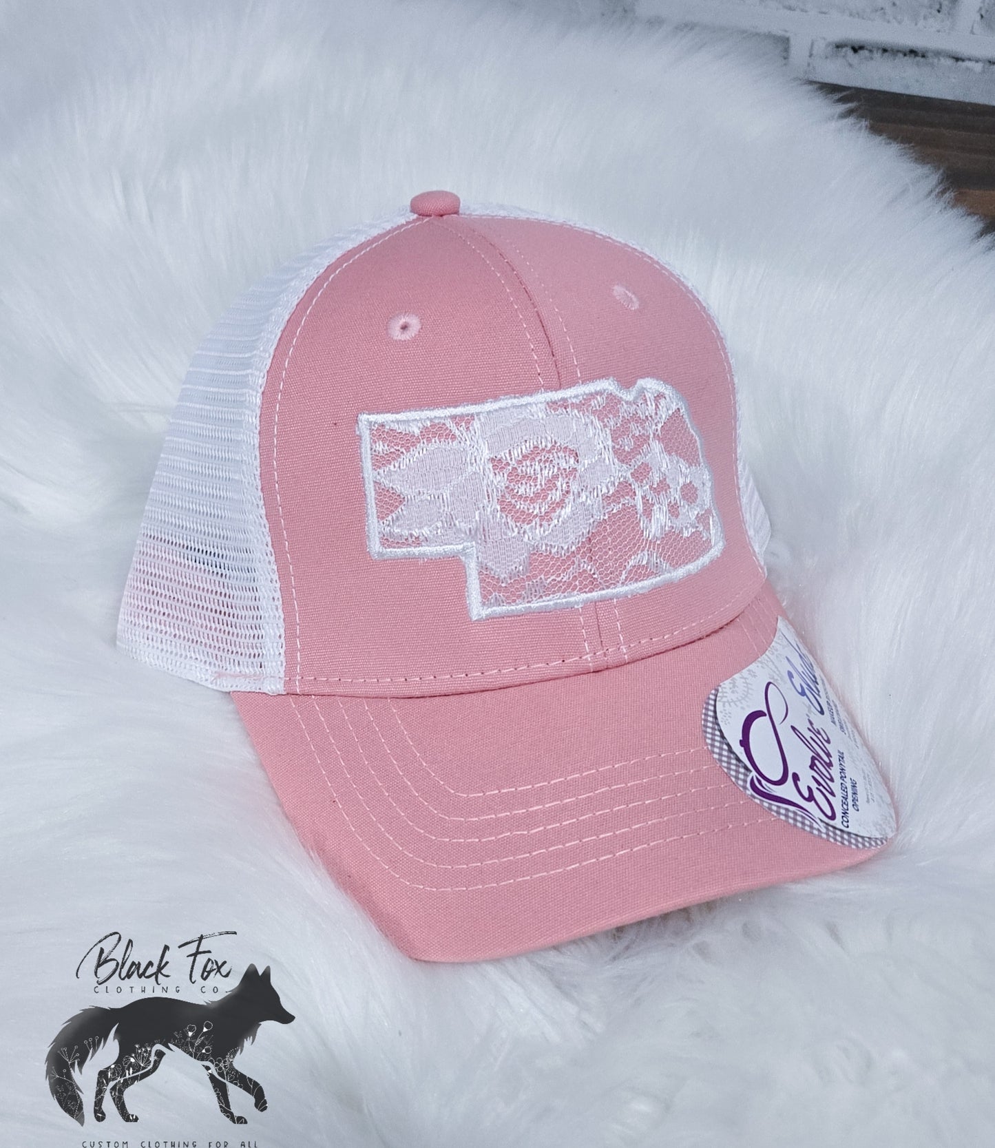 A pink Nebraska Lace applique Trucker Hat displayed on a white surface, featuring a white mesh back and a state outline design on the front.