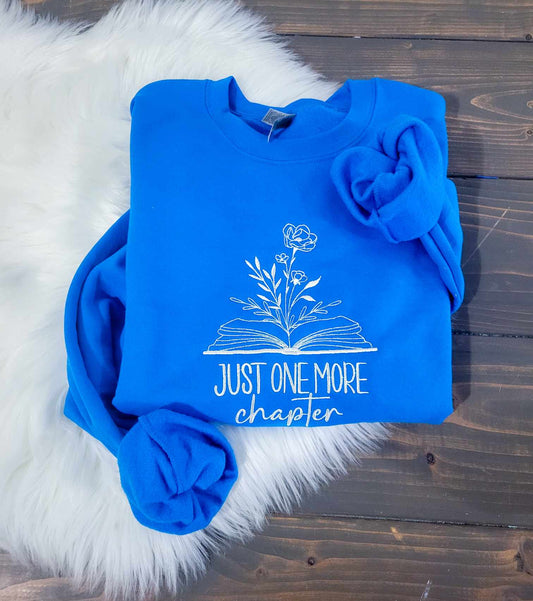 A blue crewneck sweatshirt with text just one more chapter under a book with a flower growing out of it placed on a wooden surface with a furry white object to the side.