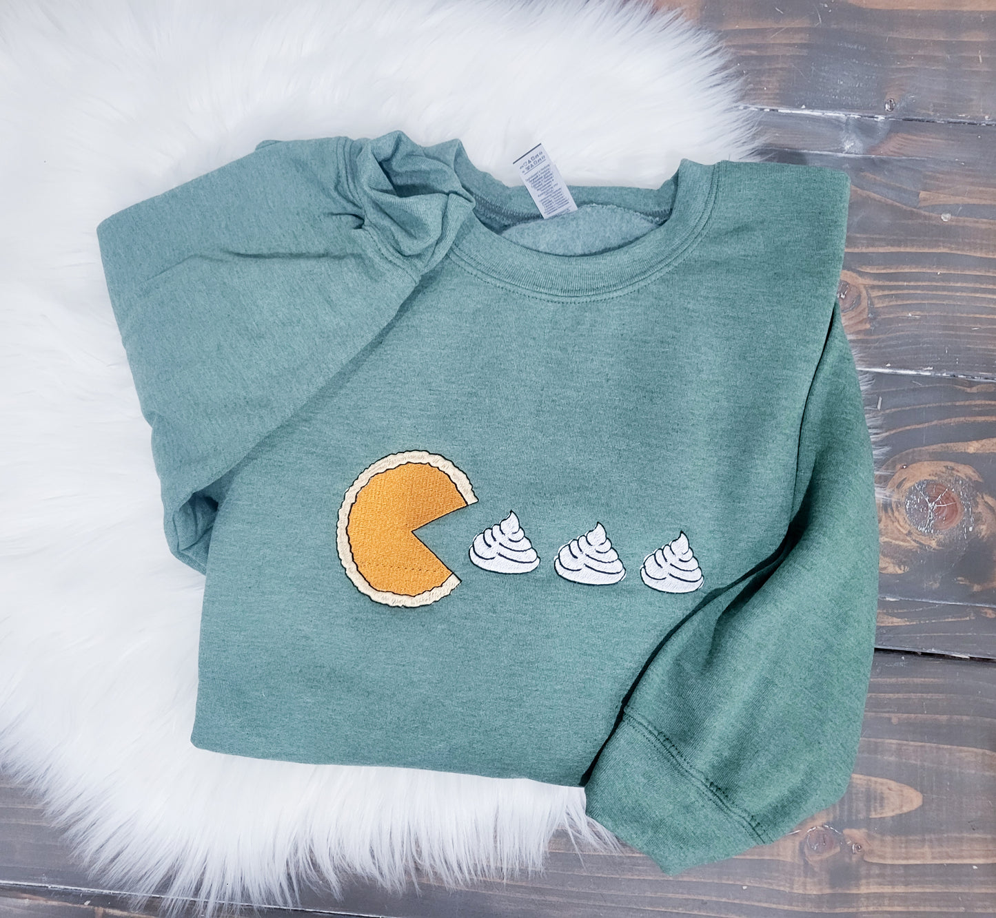 A green crewneck sweatshirt with an embroidered design of a pumpkin pie and pacman-style whip creams on a wooden surface.