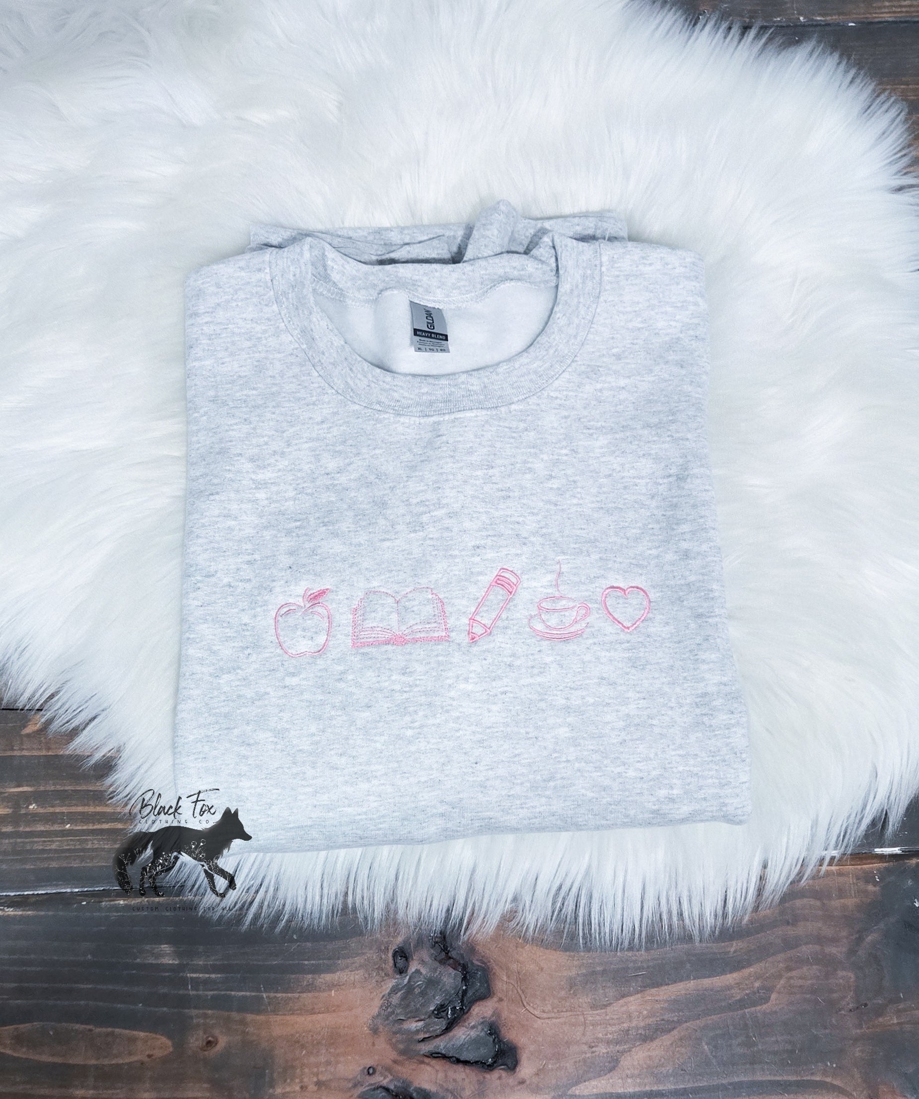 A gray fleece sweatshirt with apple, book, pencil, coffee cup and a heart embroidered  in a line on the front, displayed on a white furry surface.