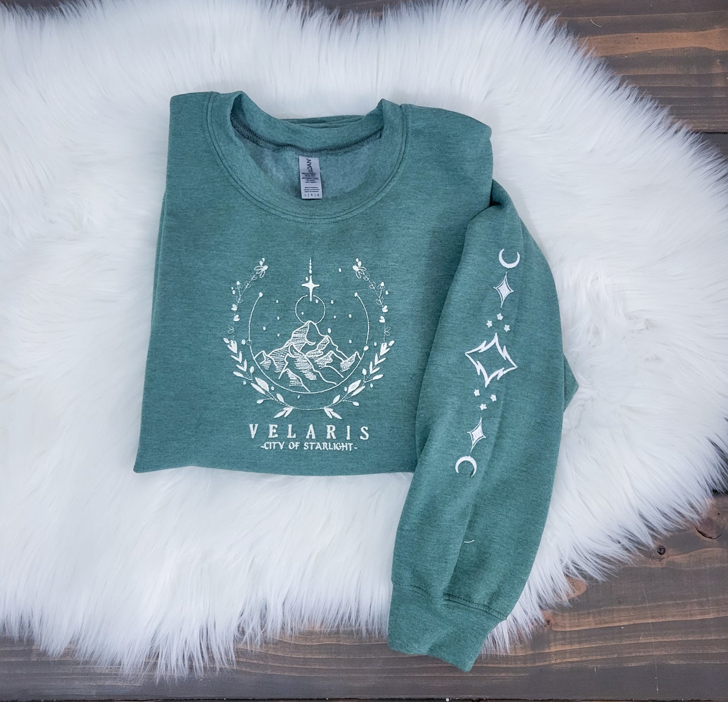 A green sweatshirt with white embroidery velaris city of starlight laid out on a white furry surface.